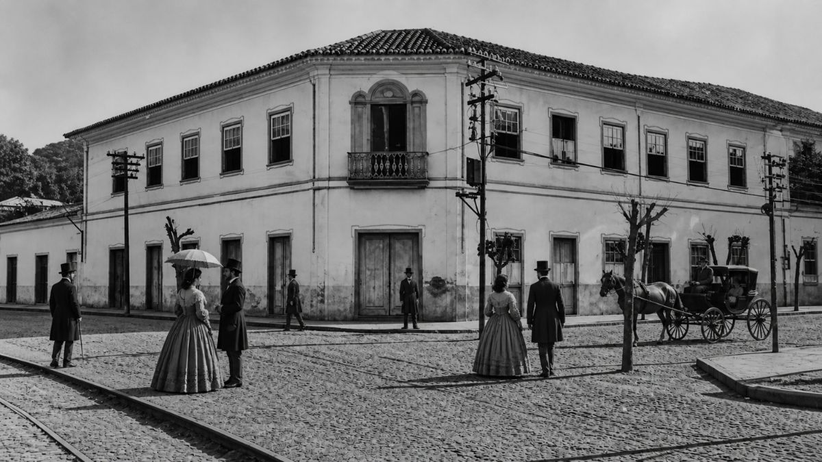 Fotografia em preto e branco mostrando um solar histórico de dois andares em uma esquina de paralelepípedos com pessoas vestidas em trajes de época e uma carruagem.
