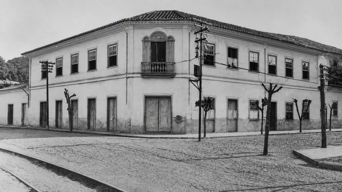 Fotografia em tons de cinza de um casarão colonial de dois andares situado em uma esquina com rua de paralelepípedos.
