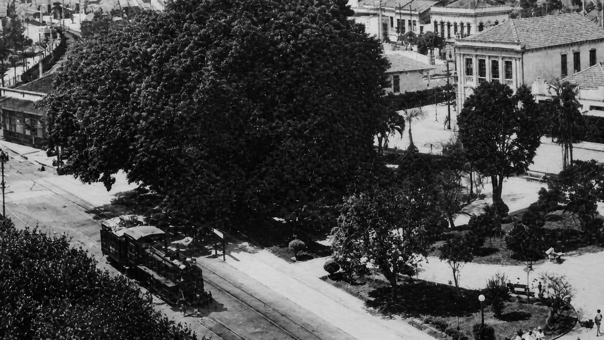Fotografia aérea em preto e branco de uma praça arborizada com trem nos trilhos.