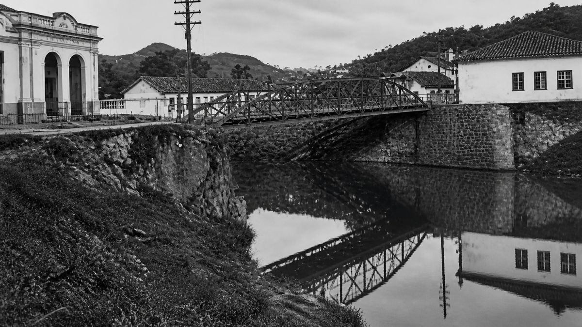 Fotografia em preto e branco mostrando uma ponte metálica sobre o Rio Piraí com construções coloniais e reflexo na água.