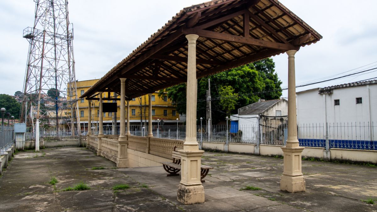 Estrutura de cobertura com telhas de barro sustentada por colunas clássicas em um pátio de cimento com bancos de madeira.