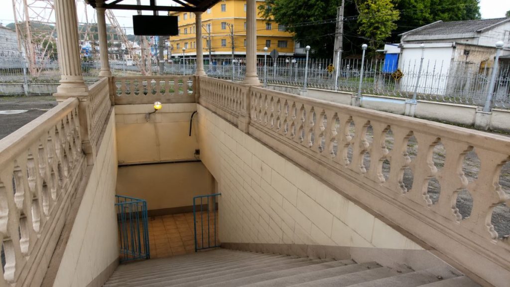 Escadaria de cimento que desce para um túnel sob um abrigo com colunas e balustrada decorativa em estilo clássico.