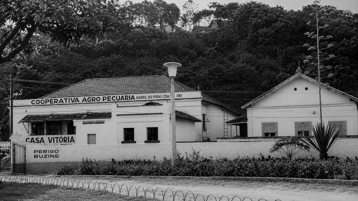 Fotografia em preto e branco de prédios antigos térreos com vegetação ao fundo.