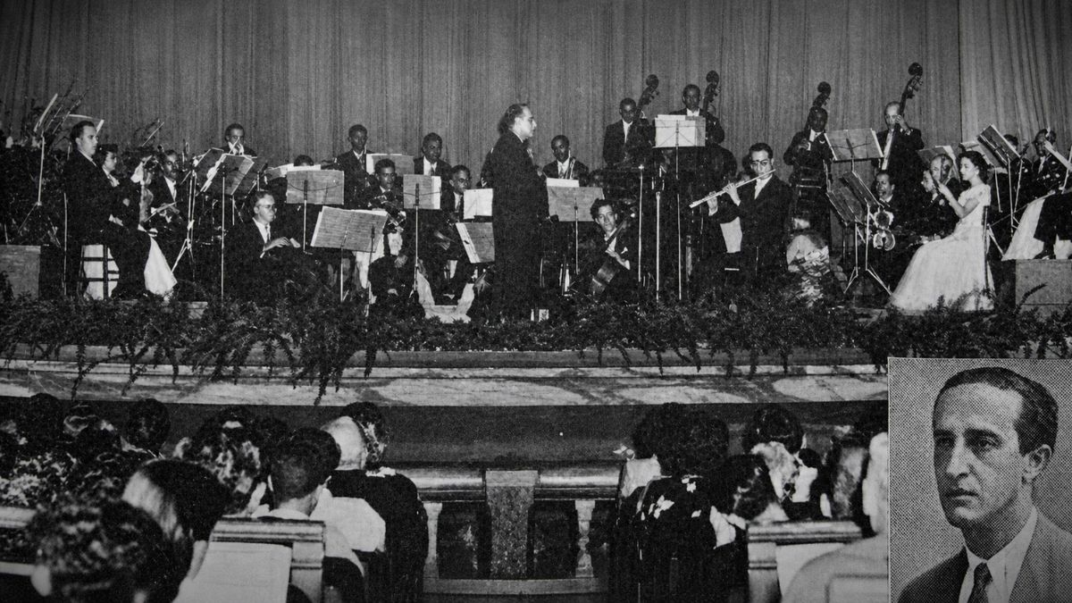 Fotografia em preto e branco de uma orquestra completa apresentando-se em um palco decorado.