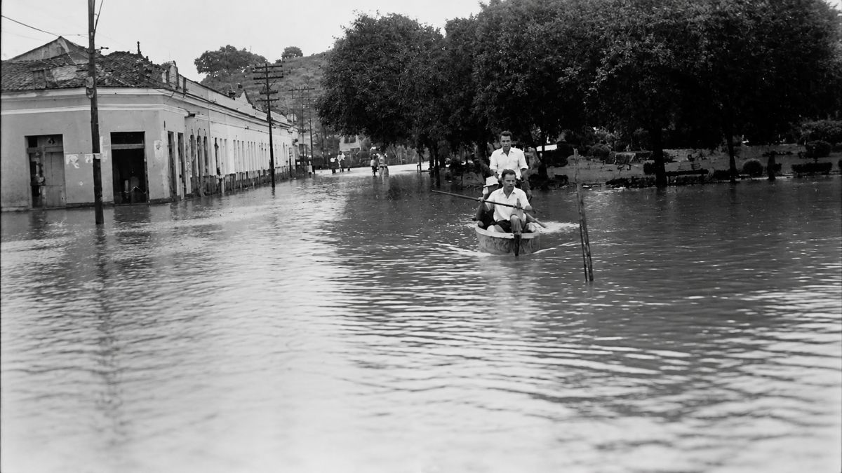 Fotografia antiga em preto e branco mostrando uma rua inundada com pessoas em uma pequena canoa.
