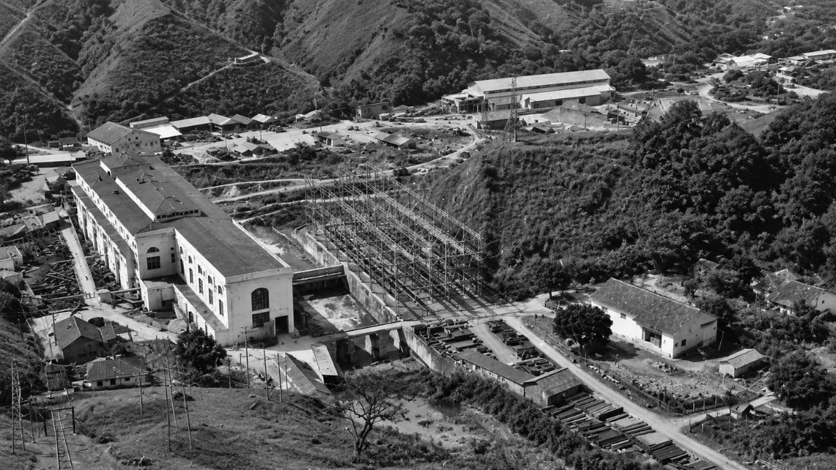 Fotografia aérea em preto e branco de um complexo industrial entre morros e vegetação.