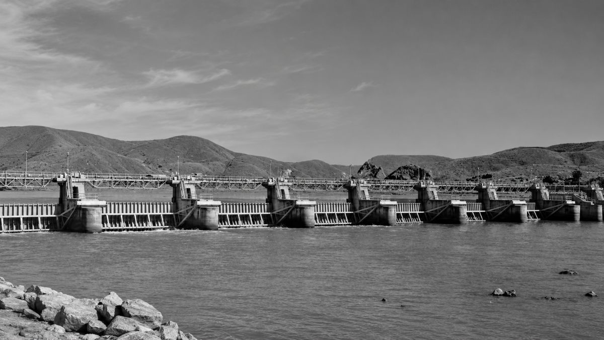 Fotografia em preto e branco de uma grande barragem hidrelétrica com comportas e montanhas.