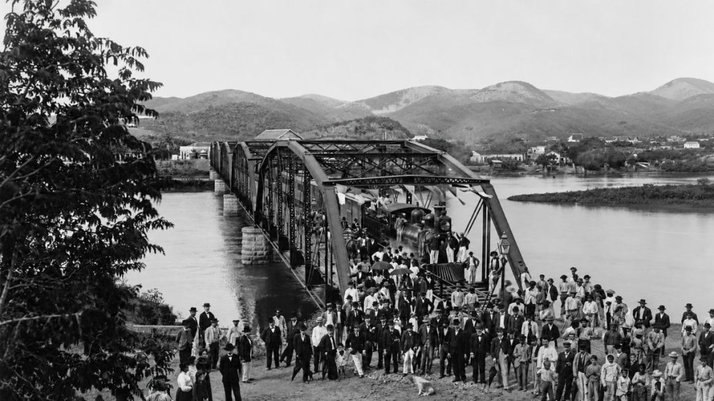 Fotografia histórica em preto e branco de uma multidão vestida formalmente celebrando a inauguração da ponte metálica com um trem.