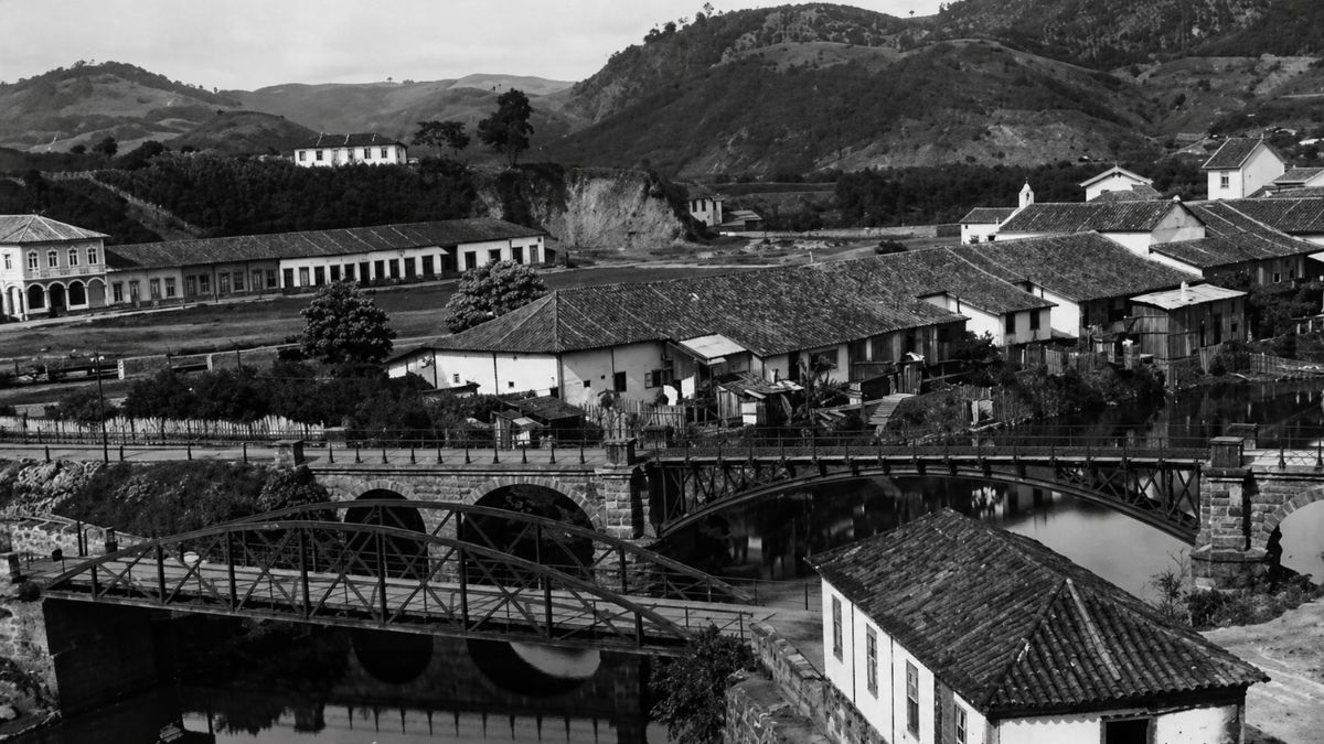 Fotografia em preto e branco de pontes metálicas sobre rio com casario e morros.