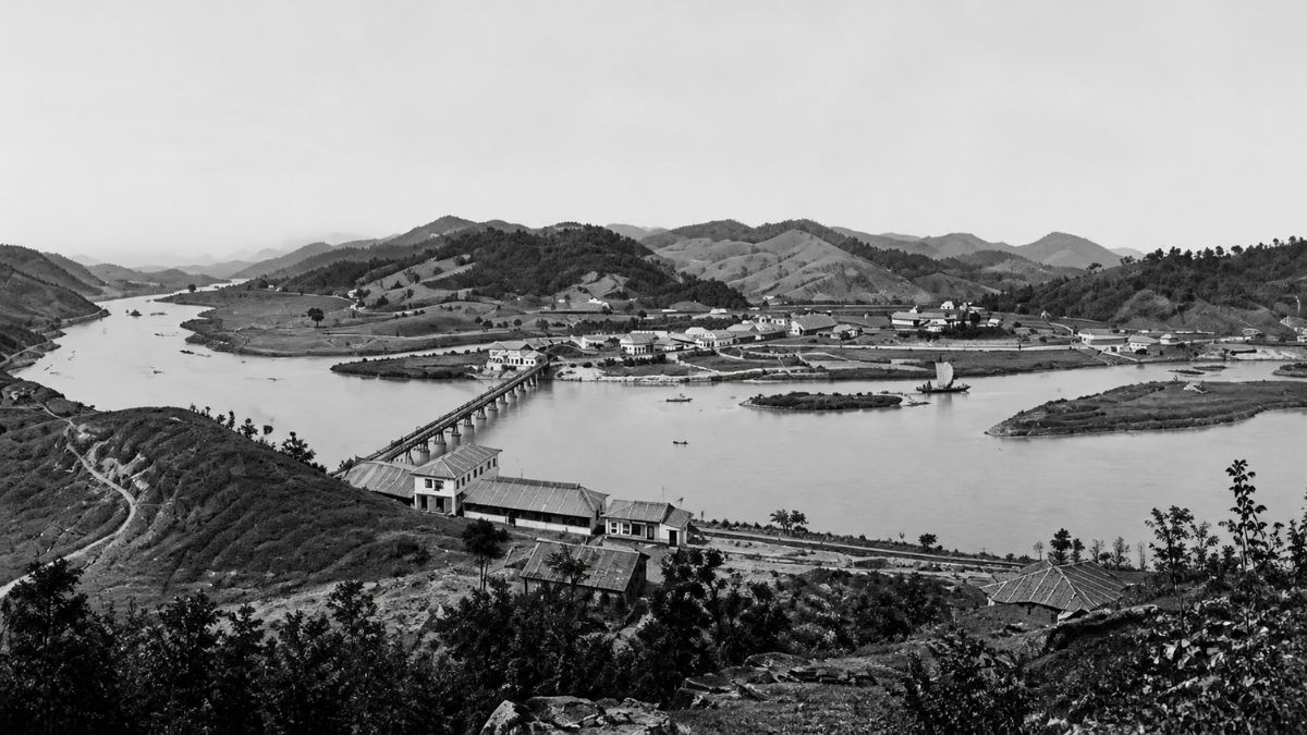 Fotografia panorâmica em preto e branco de uma vila antiga com ponte longa sobre o rio.