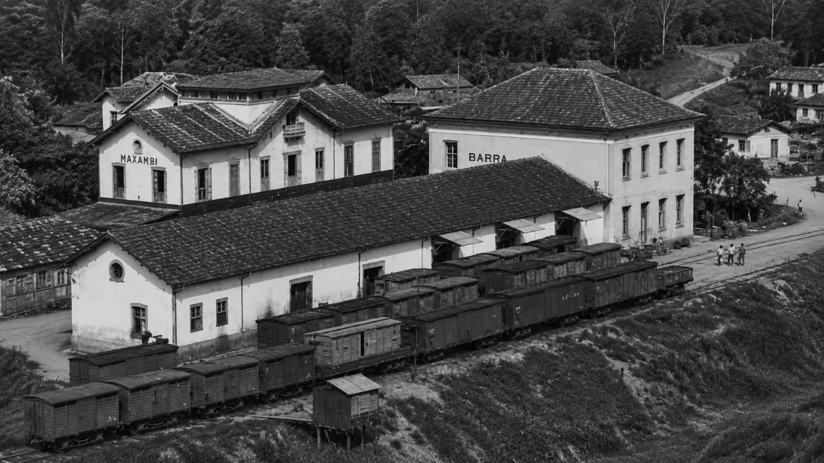 Fotografia em preto e branco de estação ferroviária com vagões de carga e prédios antigos.