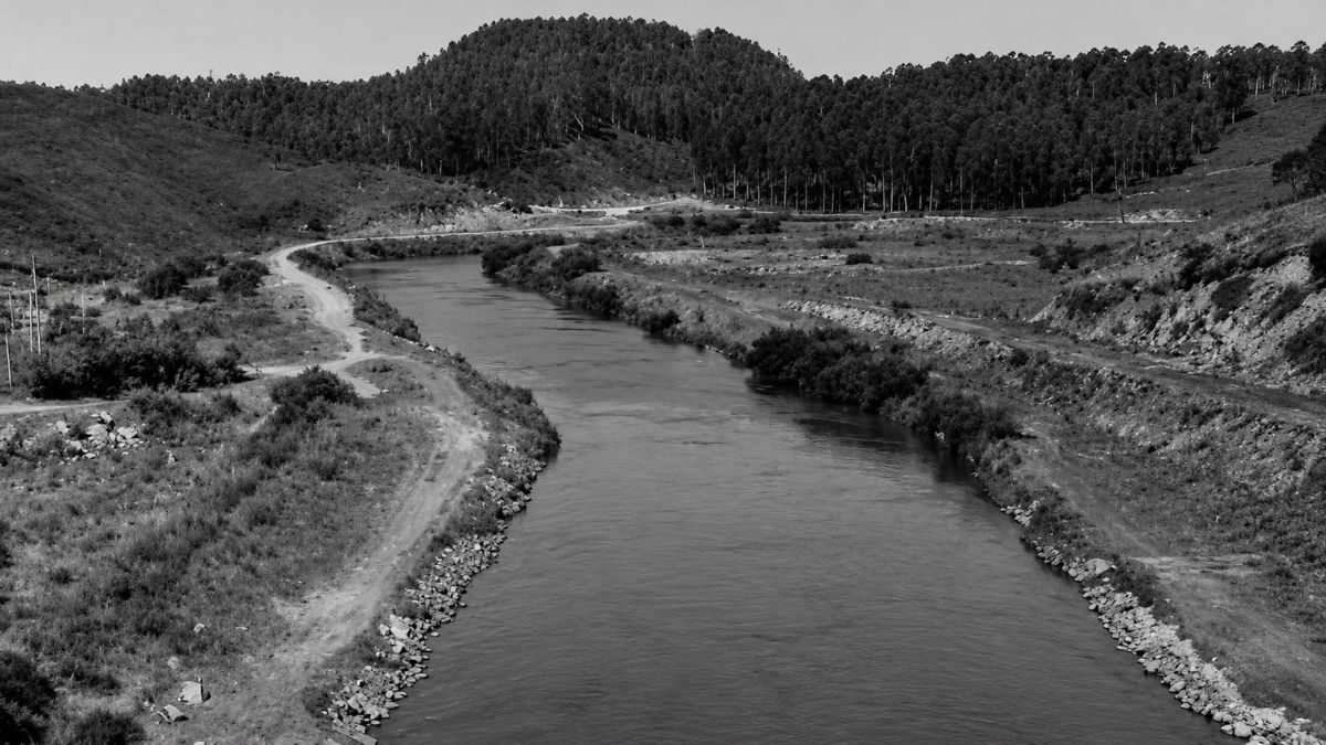 Fotografia em preto e branco de um canal largo com margens pedregosas entre morros arborizados.