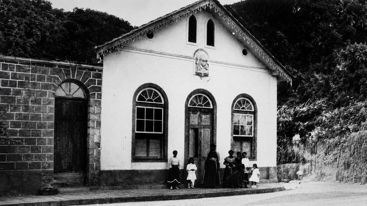 Fotografia em preto e branco de pessoas paradas diante de uma casa branca de estilo colonial.