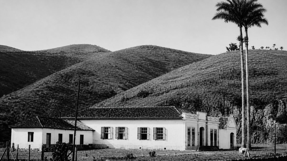 Fotografia em preto e branco de prédio colonial térreo sob morros com palmeiras ao lado.