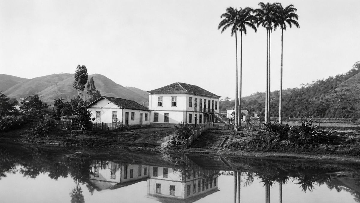 Fotografia em preto e branco de um casarão colonial refletido nas águas calmas de um rio.