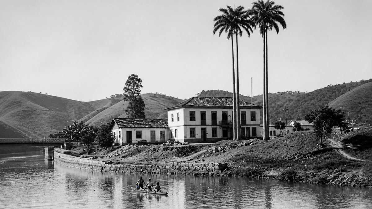 Fotografia em preto e branco de casarão colonial com palmeiras e homens em canoa no rio.