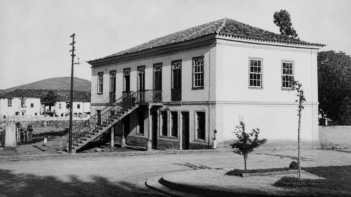 Fotografia em preto e branco de casarão colonial de dois andares com escadaria externa lateral.
