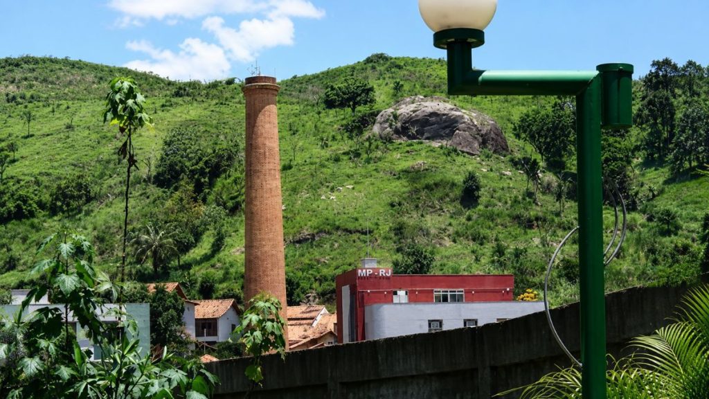 Vista da alta chaminé de tijolos ao lado de um prédio vermelho com a sigla MP RJ sob colina verde.