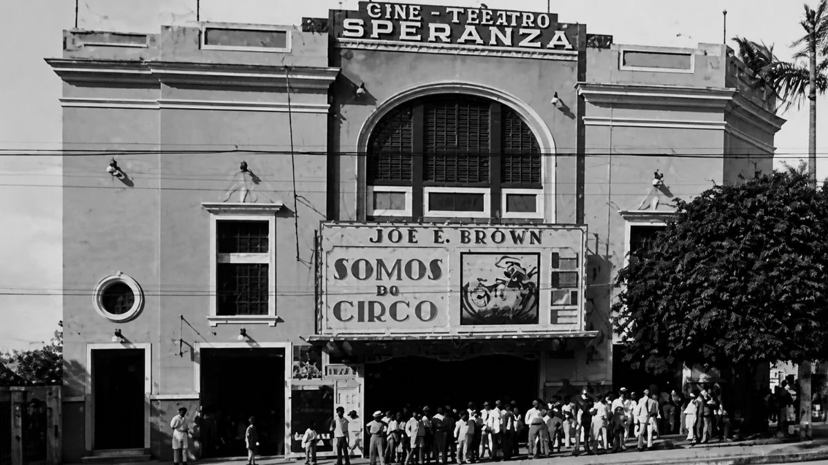 Fotografia em preto e branco de cinema antigo com multidão na porta e cartaz de filme.