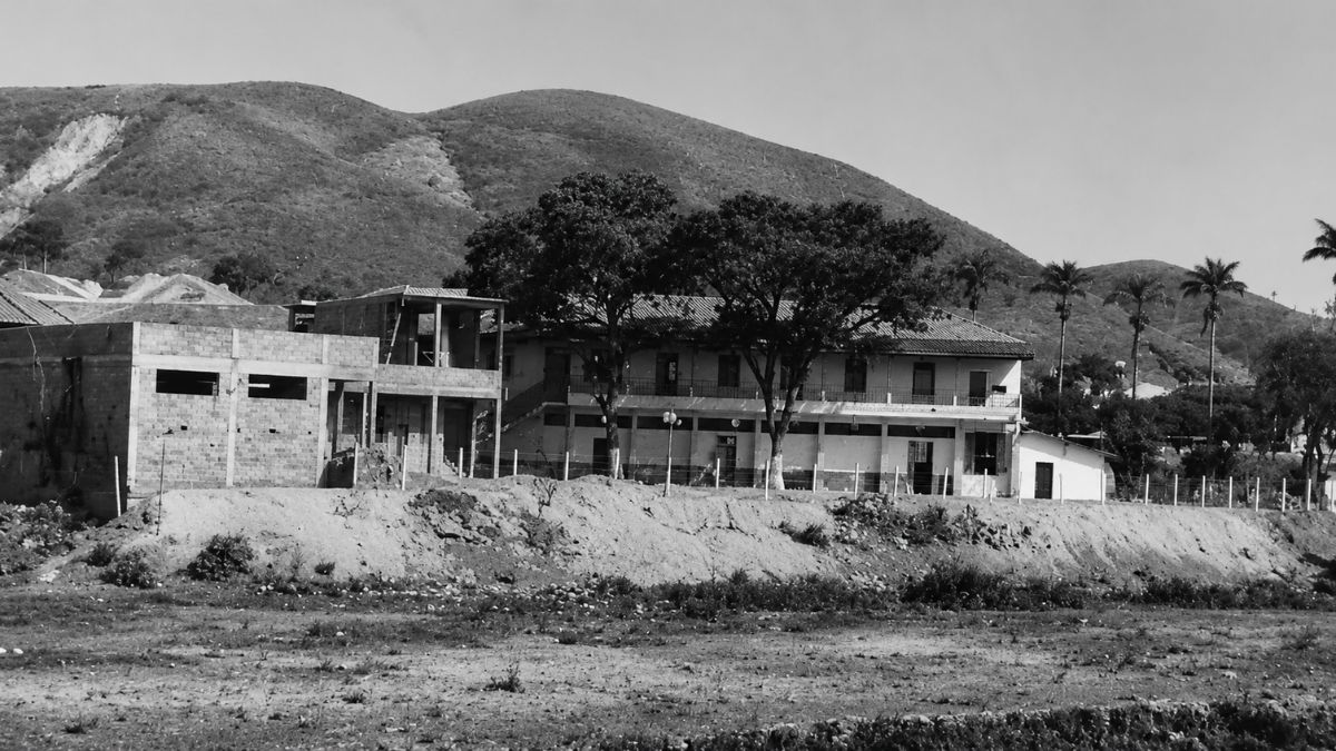 Fotografia em preto e branco de prédio escolar em obras com árvores e montanhas rasteiras.