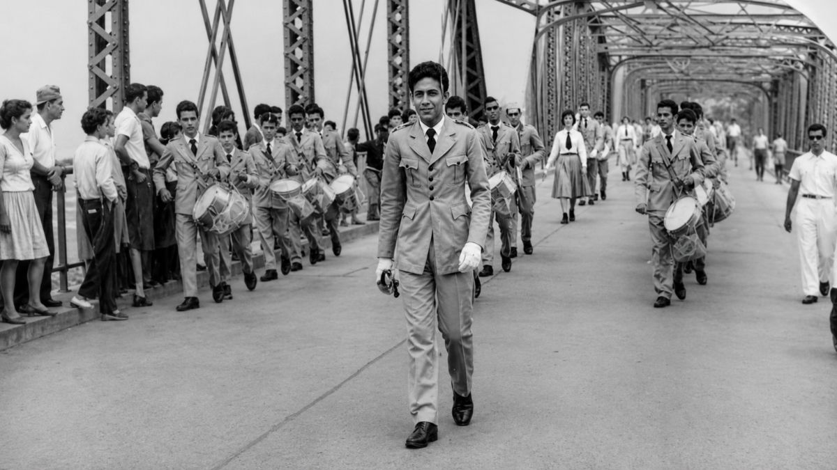Fotografia em preto e branco de estudantes fardados desfilando com tambores sobre uma ponte metálica.