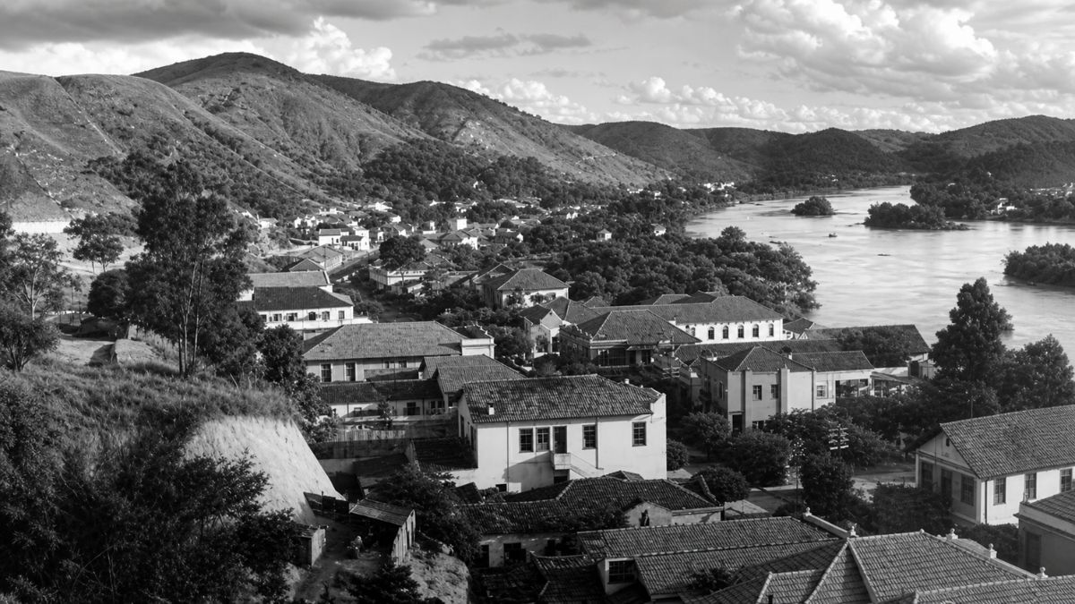 Fotografia em preto e branco mostrando casas antigas, montanhas e o curso de um rio.
