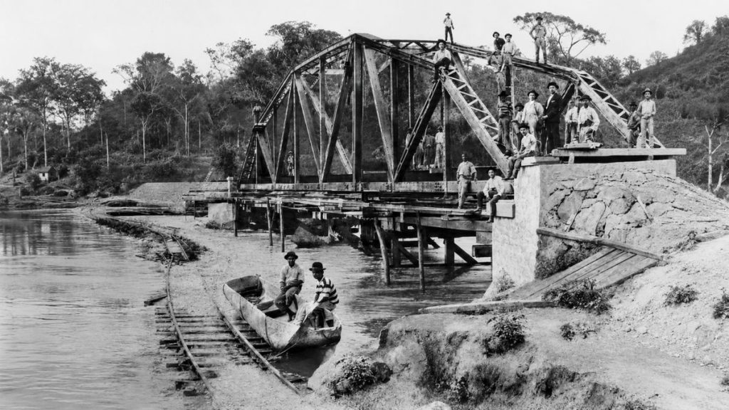 Fotografia histórica em preto e branco de operários posando sobre e ao redor de uma treliça metálica em construção.