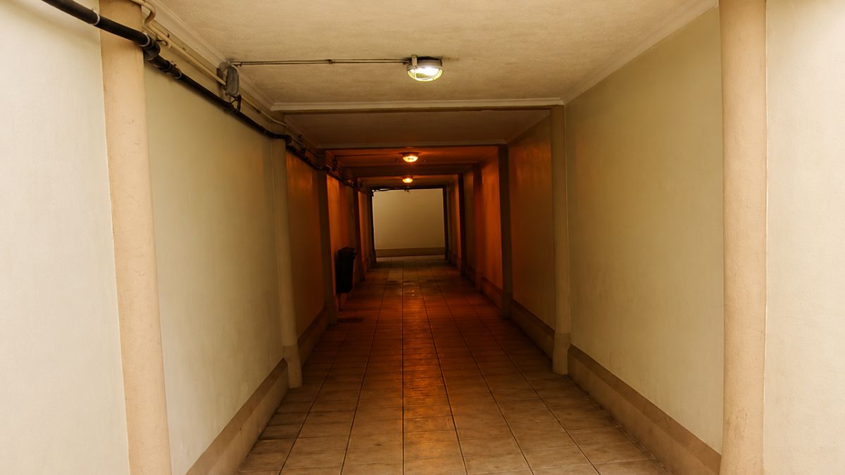 Vista interna de um túnel longo e estreito com iluminação amarelada no teto e paredes claras com colunas de suporte.