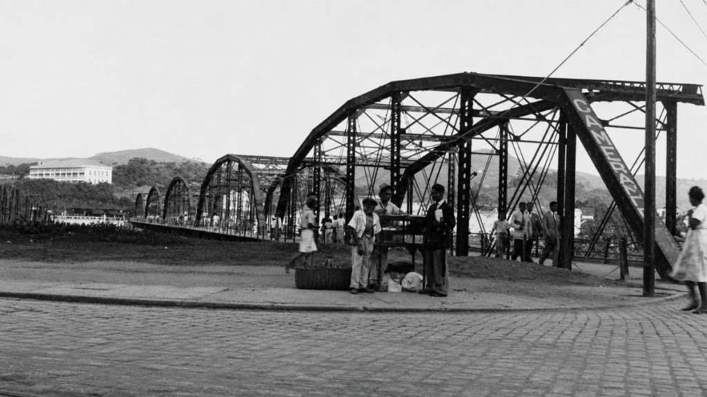 Fotografia antiga em preto e branco de uma ponte de ferro com arcos e pessoas paradas em uma rua calçada.