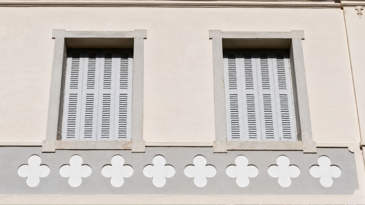 Close de duas janelas de madeira cinza com venezianas sobre uma faixa decorativa branca e cinza com desenhos de quadrifólios.