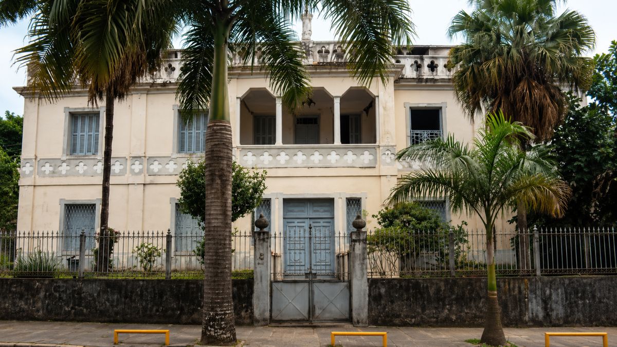 Fachada frontal centralizada de palácio bege com dois pavimentos, varanda superior com colunas, portão de ferro e palmeiras laterais.