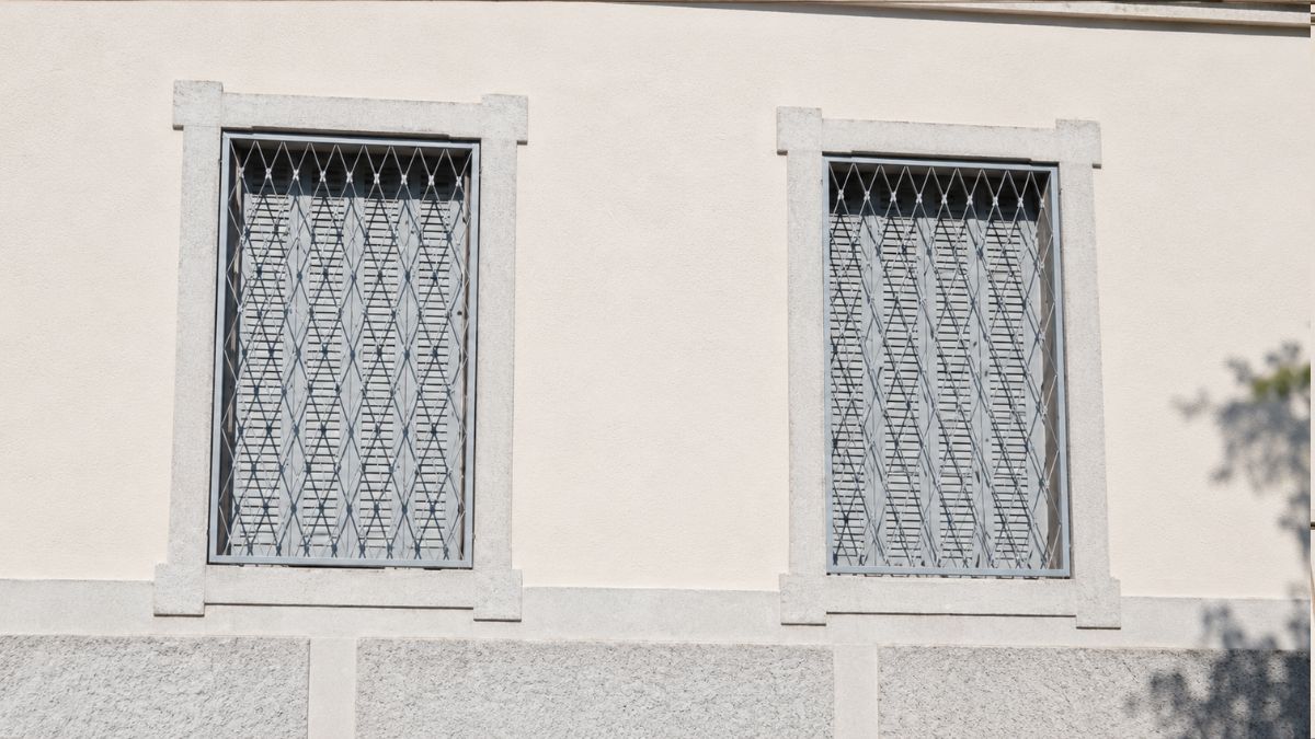 Close de duas janelas cinzas com venezianas e grades externas em formato de losangos sobre parede clara com molduras decorativas.