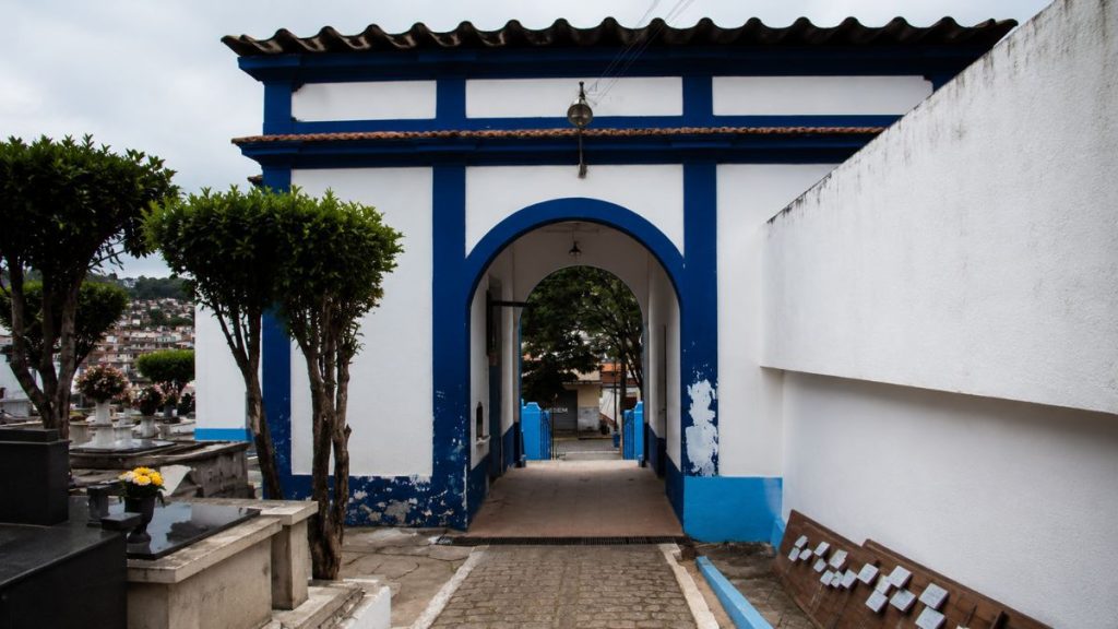 Fotografia aproximada de um pórtico branco com molduras azuis apresentando uma passagem central em arco e cobertura de telhas cerâmicas.