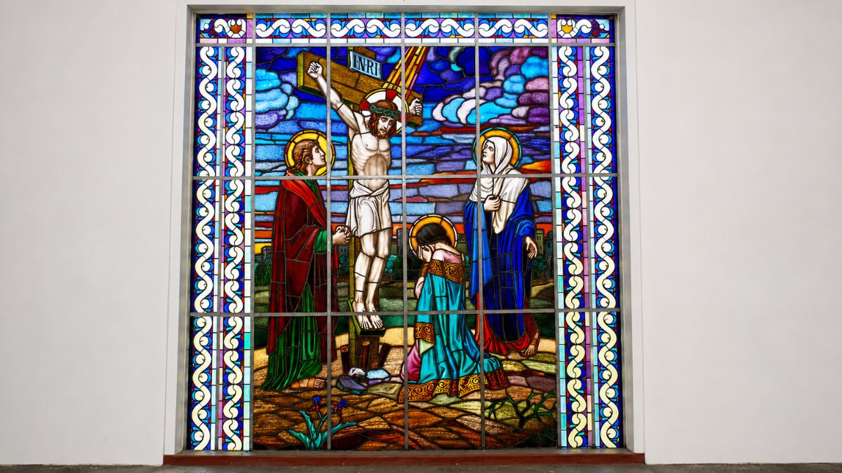 Vitral colorido em moldura quadrada representando Jesus Cristo na cruz cercado por Maria Madalena, a Virgem Maria e João.