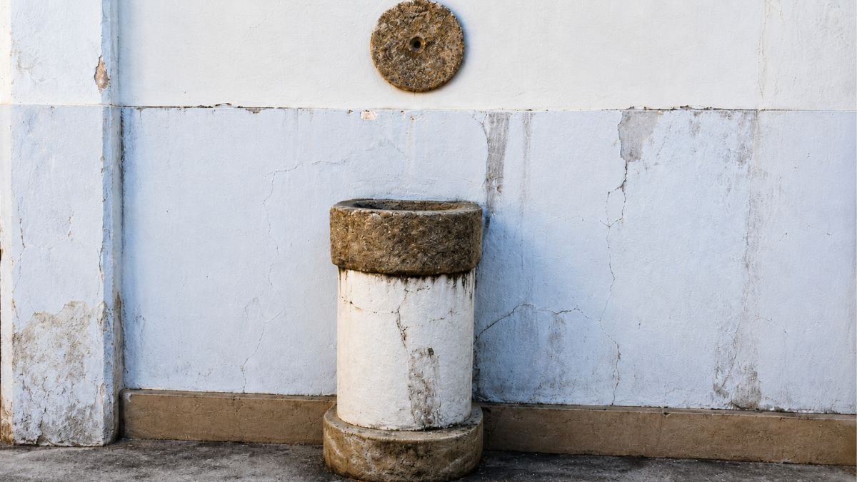 Close em uma parede azul clara com uma medalha circular de pedra acima de uma pequena coluna cilíndrica branca e rústica.