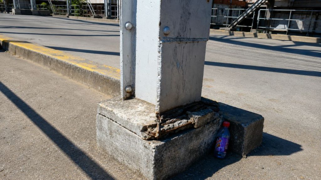 Close de base de concreto quebrada com ferragens expostas sustentando viga metálica cinza com garrafa plástica descartada ao lado.