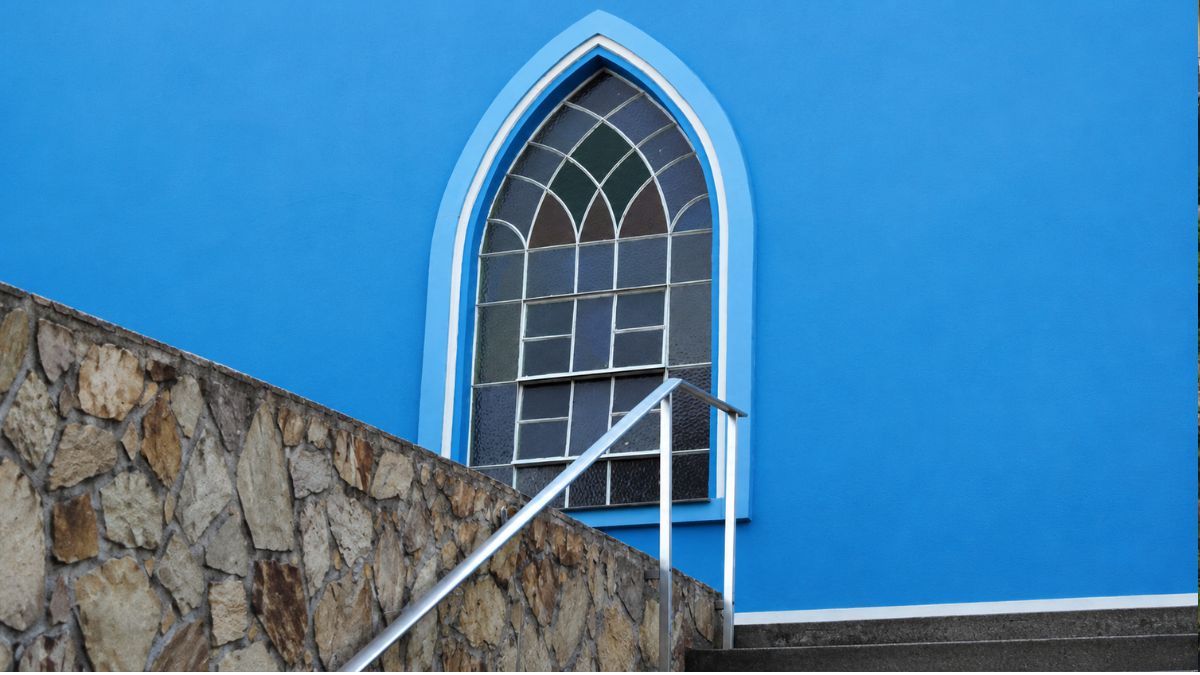 Plano detalhado de uma janela em arco ogival com vitrais e moldura branca em parede azul. À frente aparecem um corrimão metálico e muro de pedra.