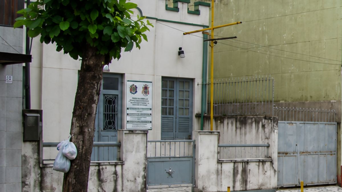 Close da entrada do prédio militar mostrando portão cinza com estrela, placa informativa na parede e parte de uma árvore.