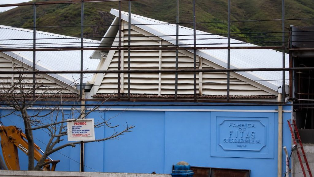 Fotografia aproximada de parede azul com placa em relevo sob telhado branco tipo shed e estrutura metálica de obra.