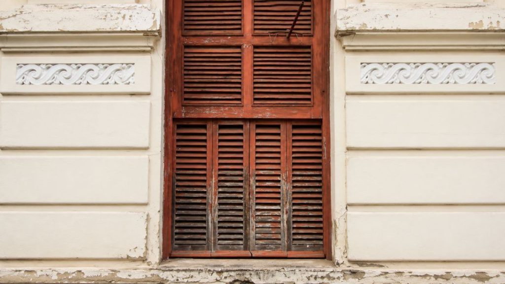 Close de uma janela de madeira avermelhada com venezianas horizontais emoldurada por paredes de tom creme com frisos decorativos.
