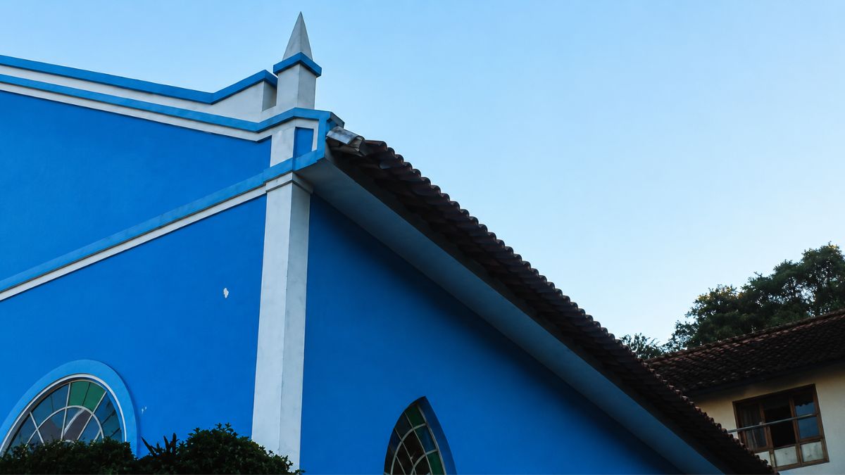 Close do ângulo superior de uma igreja azul mostrando o beiral do telhado cerâmico, um pináculo branco e uma platibanda. Parte de uma janela em arco ogival aparece abaixo.
