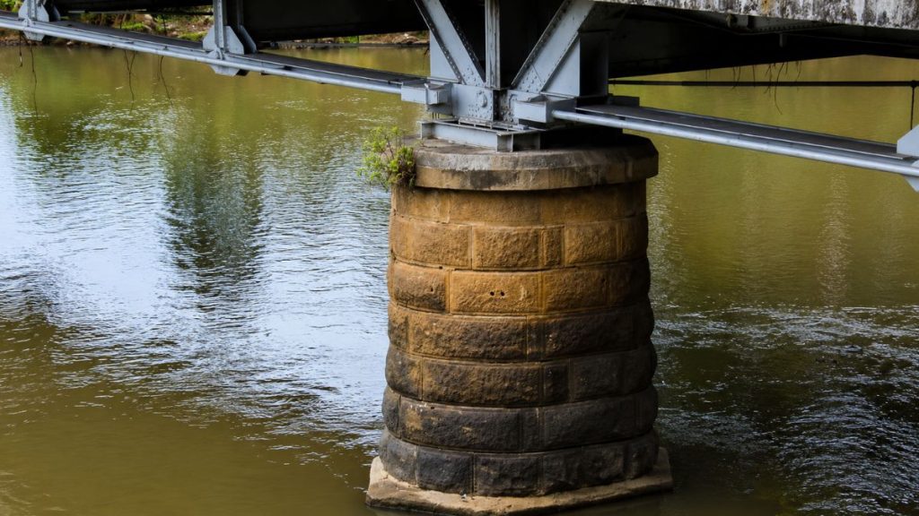 Pilar cilíndrico de alvenaria de pedra sob estrutura metálica da ponte mergulhado em águas turvas de rio esverdeado.