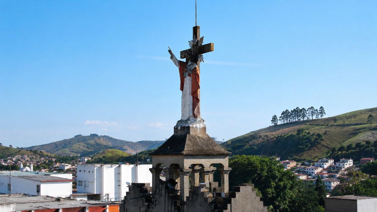 Fotografia em close do topo de uma torre escalonada com escultura de Jesus Cristo em frente a uma cruz de pedra.