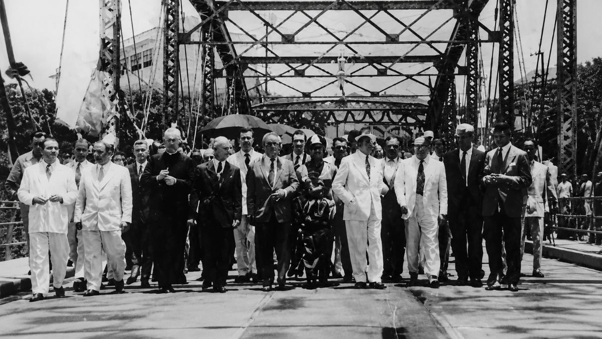 Fotografia em preto e branco de homens de terno caminhando sobre uma ponte metálica.