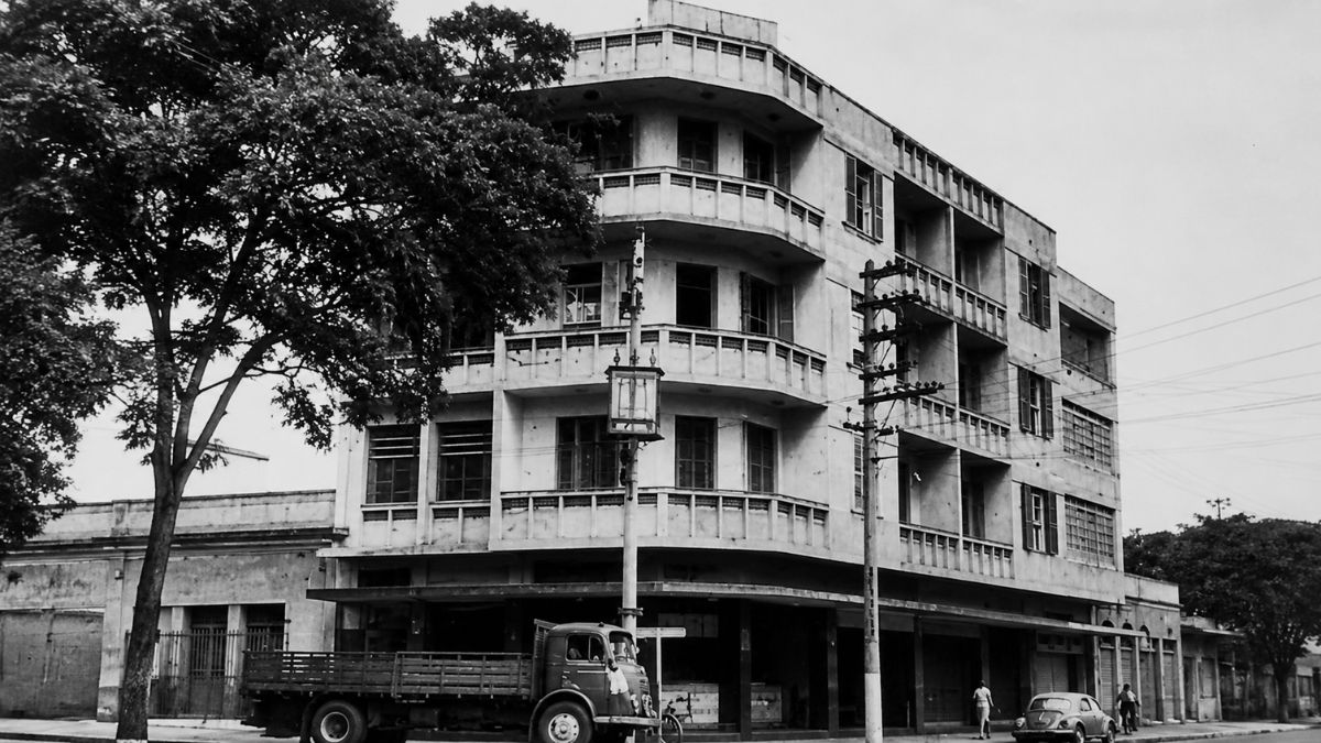 Fotografia em preto e branco de prédio de esquina com sacadas e veículos antigos.