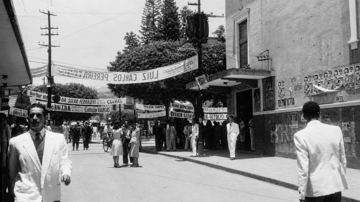 Fotografia em preto e branco de rua movimentada com faixas políticas e pessoas trajadas.