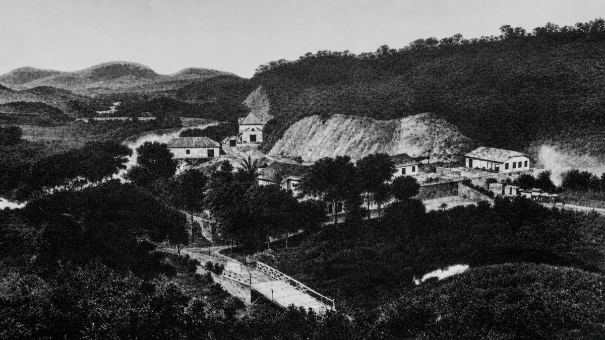 Fotografia antiga em preto e branco mostrando pequenas edificações rurais entre morros e árvores.