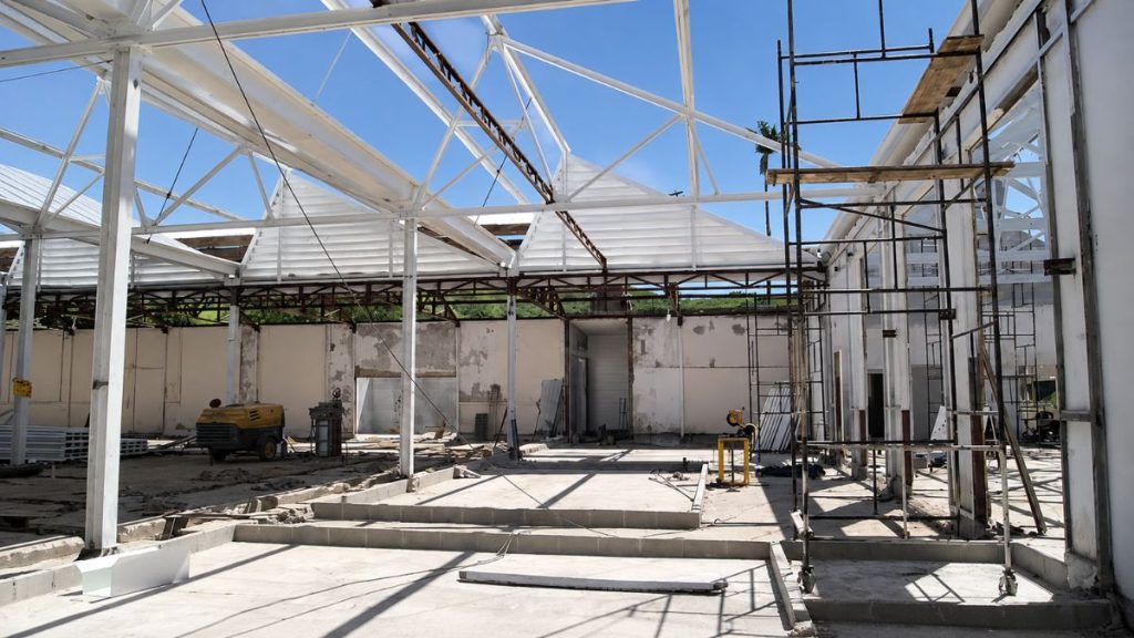 Fotografia do interior da fábrica em obras com vigas metálicas brancas aparentes, andaimes, piso de concreto e céu aberto.