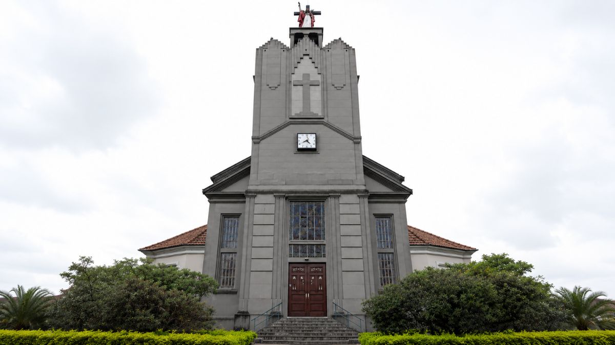 Fotografia frontal de igreja art déco cinza com torre central escalonada, vitral sobre a porta principal e estátua no topo.