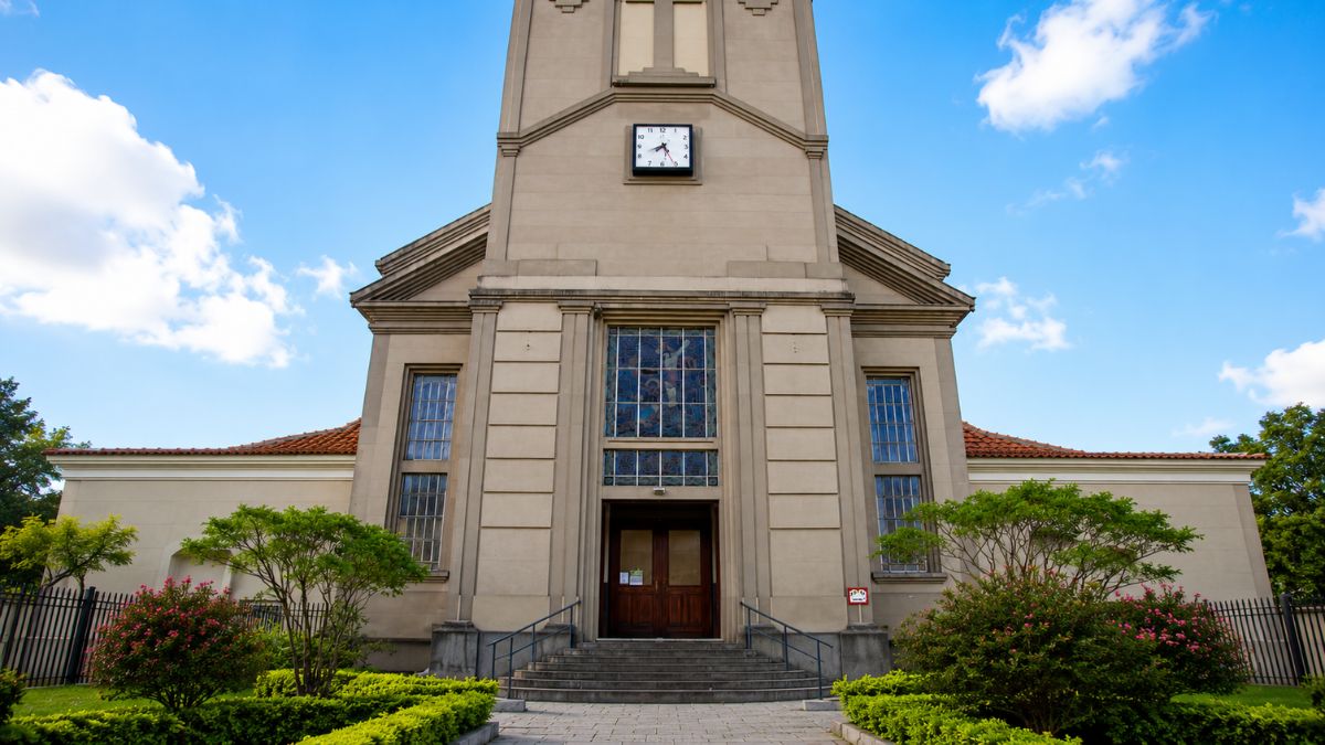 Fotografia em perspectiva frontal da entrada de uma igreja art déco cinza com escadaria, porta dupla de madeira, vitral colorido, relógio e torre